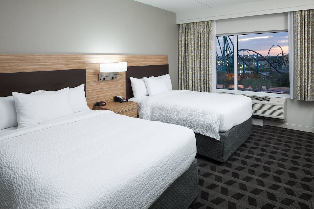 A hotel room with two beds, a nightstand, a lamp, a window with a view of a roller coaster, and a patterned carpet.