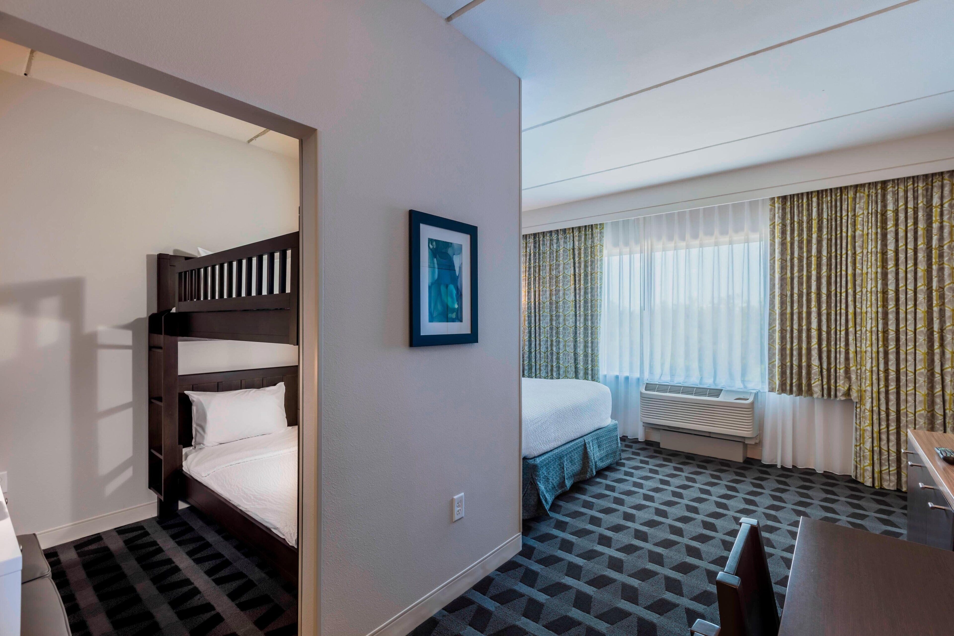 A hotel room with a bunk bed, a single bed, a desk, and a window with curtains.