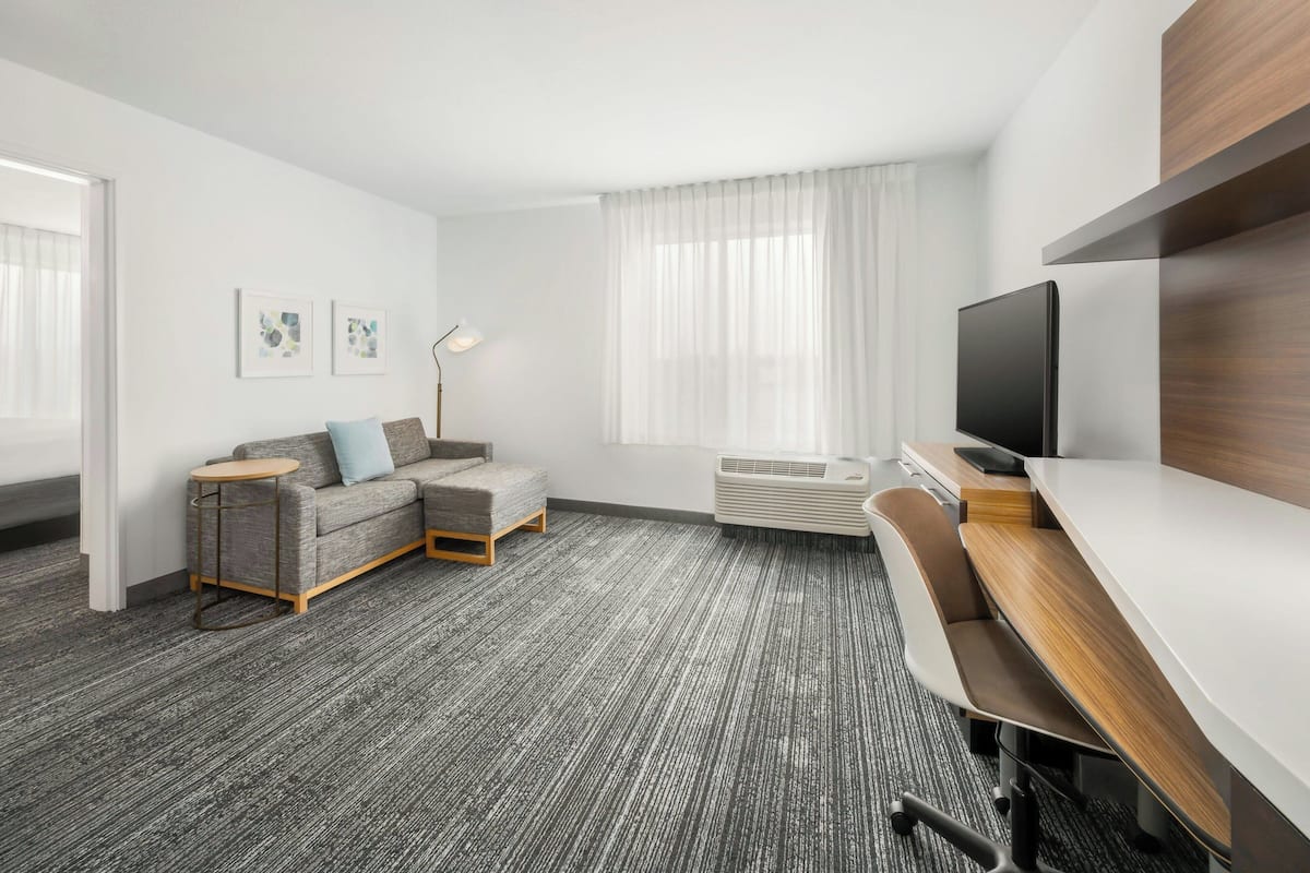 Een moderne hotelkamer met een grijs tapijt, een grijze bank met een klein rond tafeltje, een televisie op een houten meubel en een groot raam met transparante gordijnen.