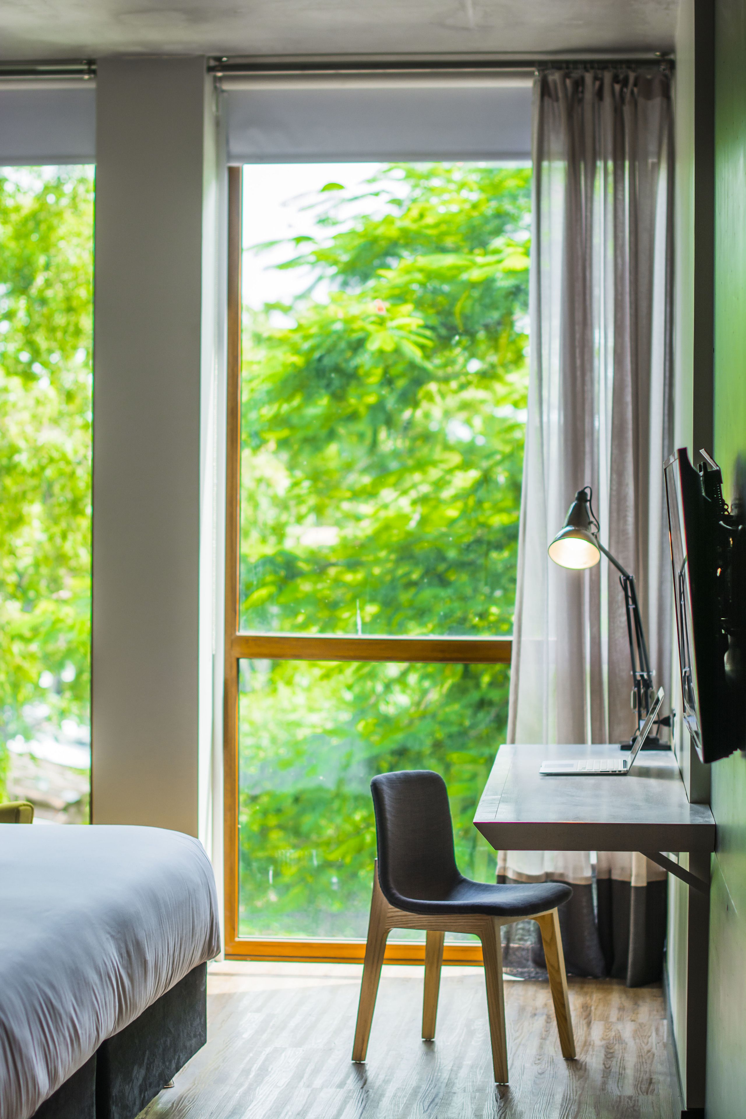A room with a bed, a desk with a chair, a lamp, and a large window with a view of green foliage.