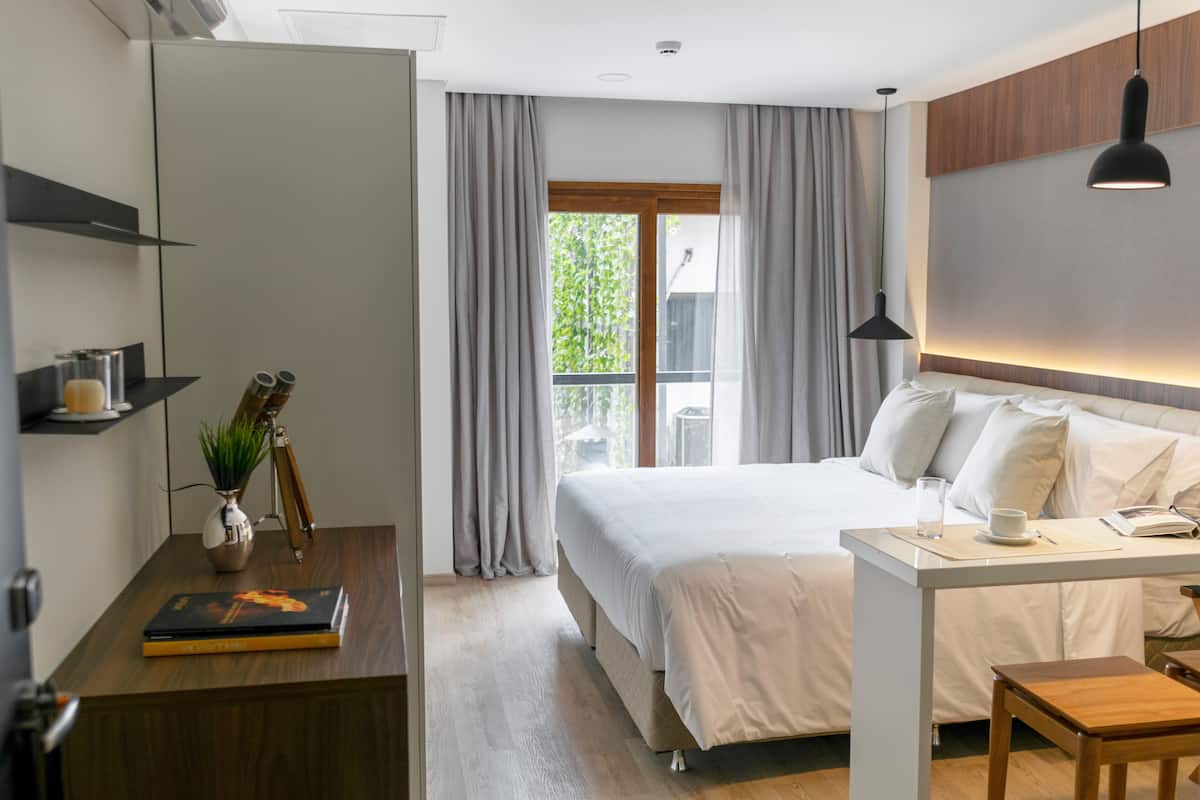 A modern hotel room with a large bed, a desk with a laptop, a small table, and a view of greenery outside.