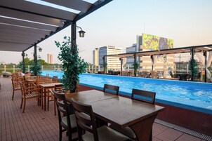 Ein Außenbereich zum Essen mit Pool, Holztischen und -stühlen sowie Stadtgebäuden im Hintergrund.