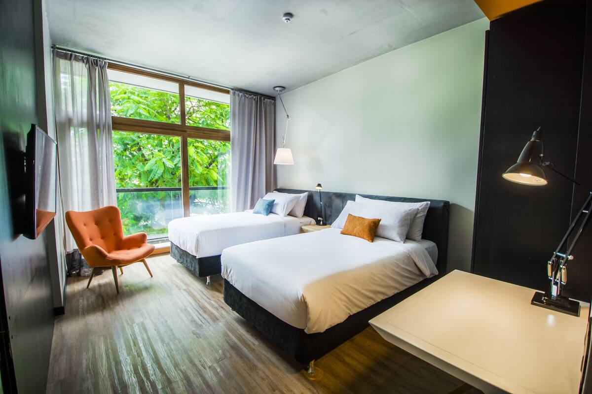 A hotel room with two beds, a chair, a desk, and a large window with curtains.