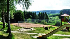 Ein Spielplatz mit Rutsche und einem eingezäunten Tennisplatz, umgeben von Bäumen und Hügeln.