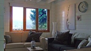 Ein gemütliches Wohnzimmer mit einem beigefarbenen Sofa, einem dunklen Ohrensessel und einem kleinen Tisch mit einer Kerze. Große Fenster bieten Blick auf viel Grün draußen.