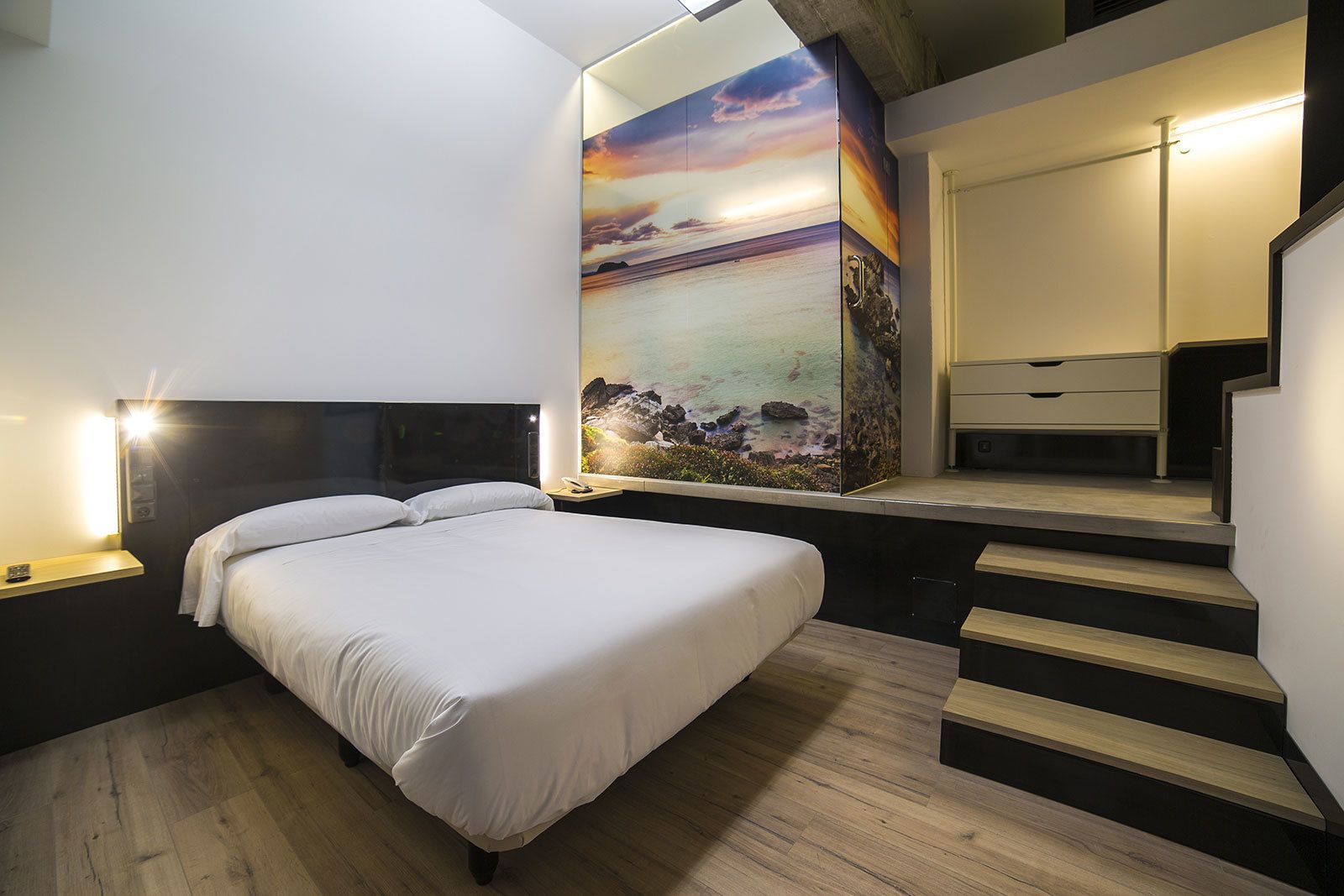 A modern bedroom with a large bed, a nightstand, a wall-mounted TV, and a staircase.