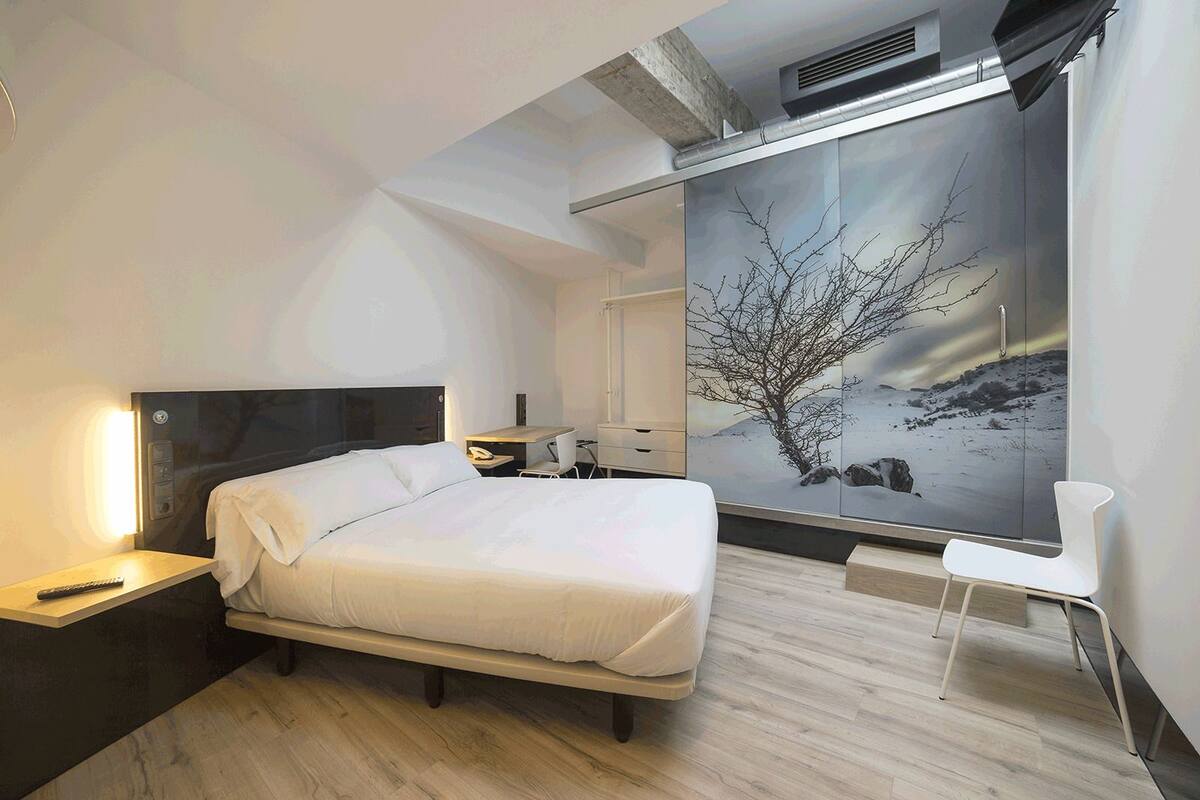 Une chambre d’hôtel moderne équipée d’un lit, d’une table de chevet, d’un bureau, d’une chaise et d’une grande fenêtre donnant sur un paysage hivernal.
