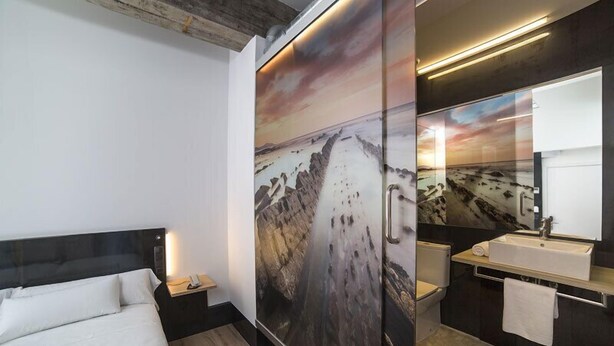 A modern hotel room with a large bed, a nightstand, a bathroom with a sink and mirror, and a scenic mural on the wall. - Hotel Zerupe (Zarautz)