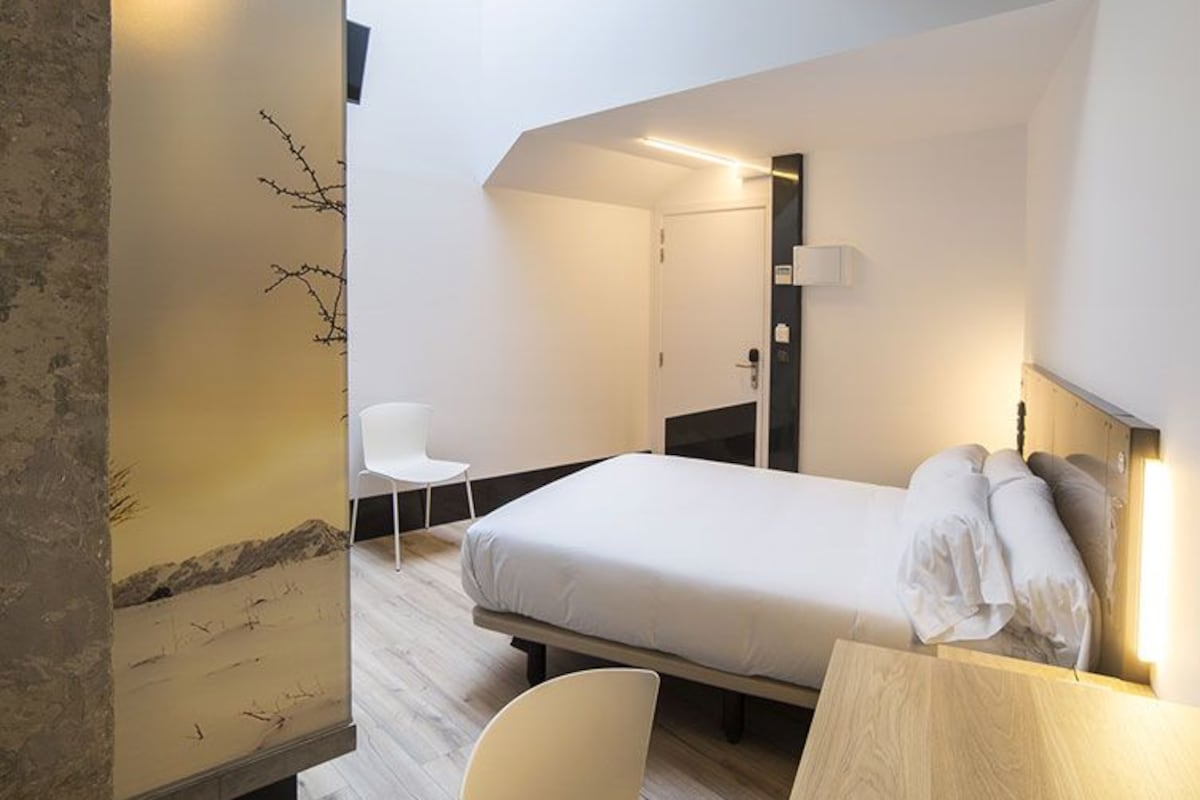 Una habitación de hotel moderna con cama, escritorio y silla. Cuenta con un amplio ventanal que proporciona luz natural y vistas al cielo.