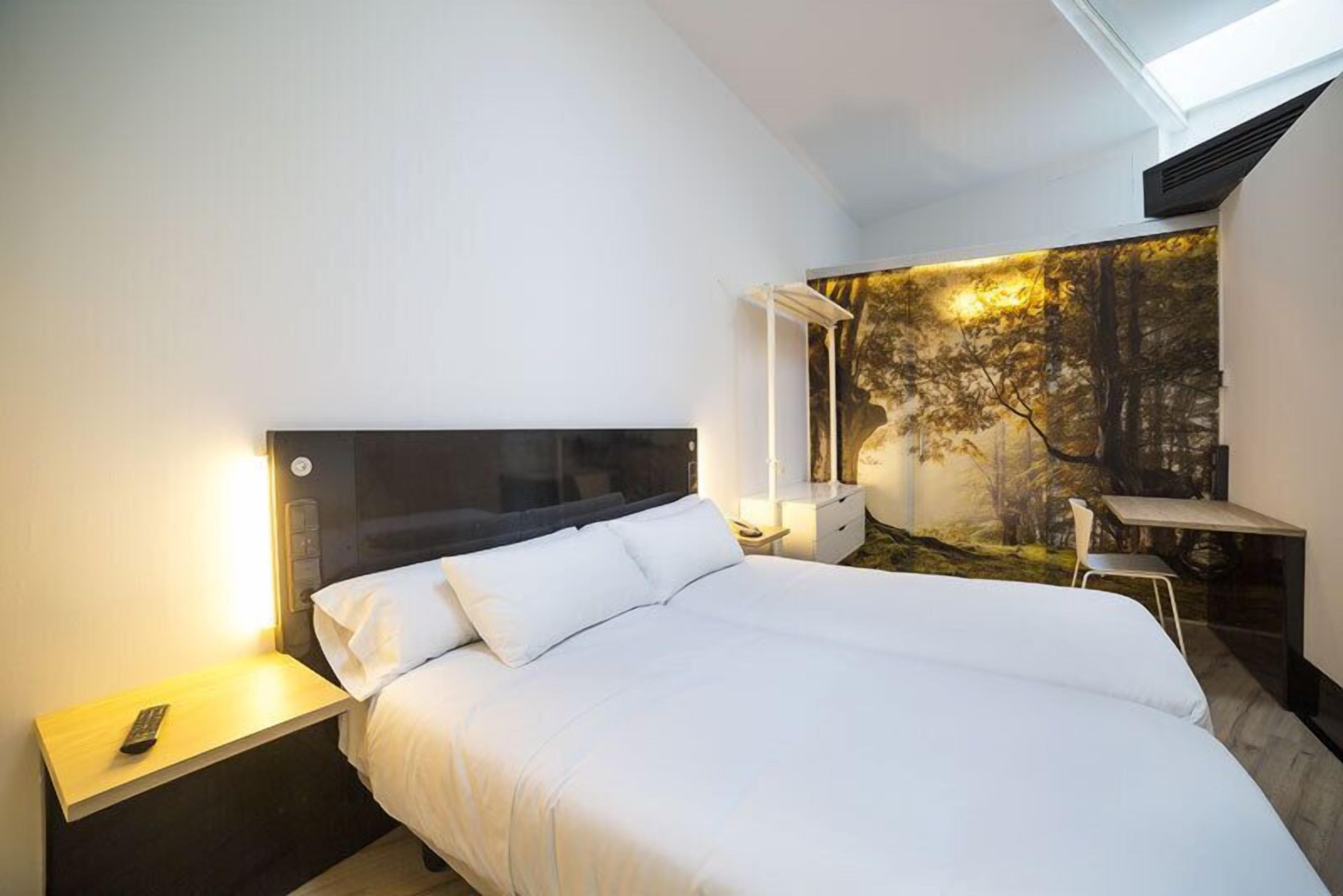 Een moderne hotelkamer met een groot bed, een nachtkastje met een afstandsbediening, een bureau met een stoel en een wanddecoratie met een bosmotief.