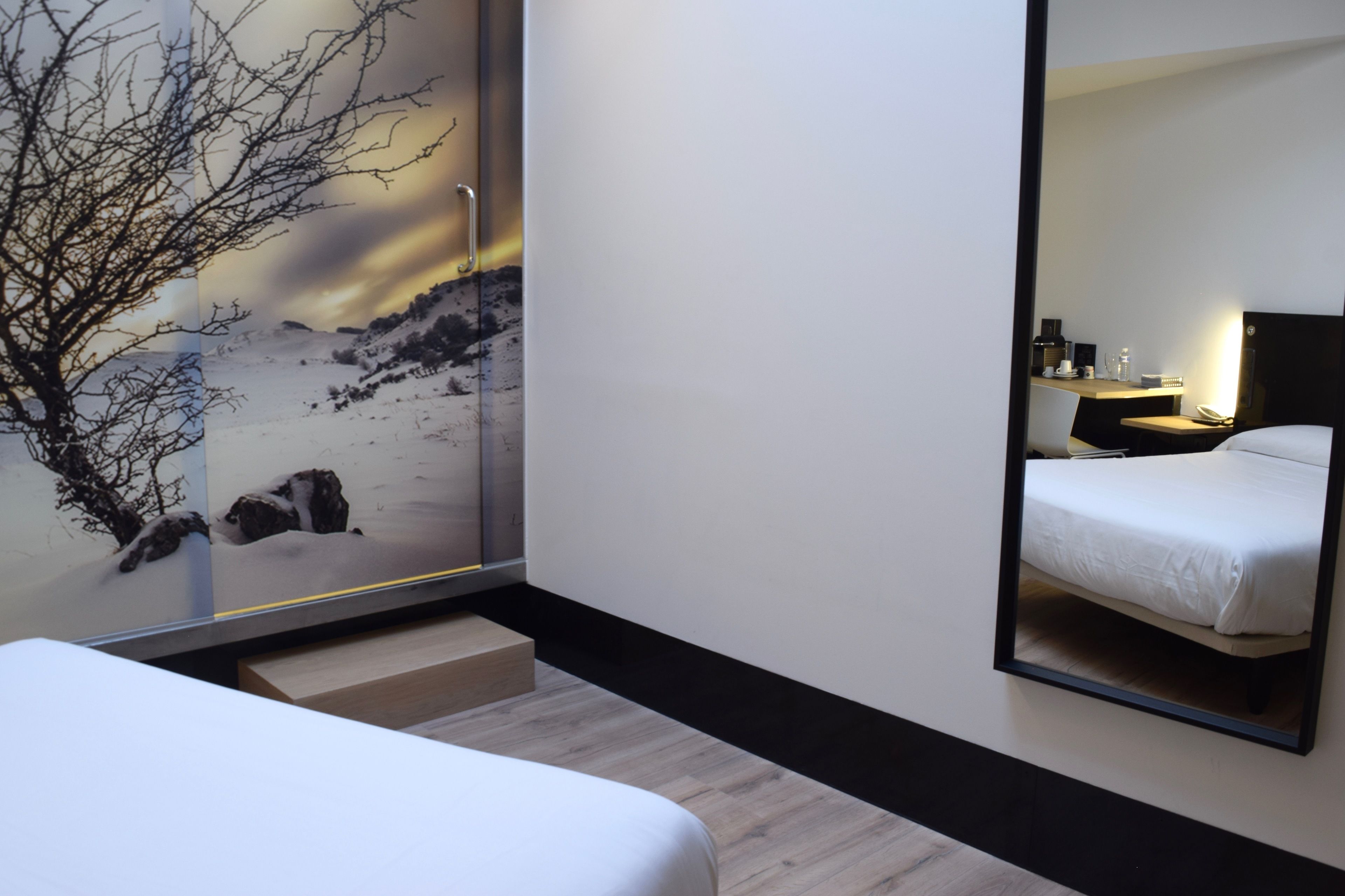 A modern hotel room with a large bed, a nightstand, a mirror, and a scenic wall mural.