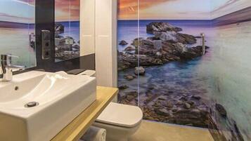 Un baño con lavamanos, inodoro y un mural con vistas a una playa rocosa.