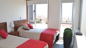A hotel room with two beds, a window with a view of buildings, a small table with a lamp, and a potted plant. - Lofts Magnolio Cerro Alegre (Valparaiso)