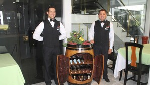 Two waiters standing next to a wine barrel display with bottles and a decorative plant. - Hotel & Resort CIDE (Minga Guazú)