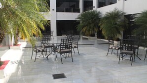 An outdoor seating area with tables and chairs, surrounded by palm trees and a modern building in the background. - Hotel & Resort CIDE (Minga Guazú)