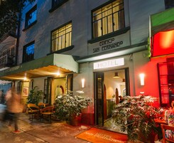 L’entrée d’un hôtel, avec une enseigne indiquant « HÔTEL SAN FERNANDO » et une porte bien visible. Des plantes en pot sont disposées à l’extérieur, et un paillasson porte le nom de l’hôtel.