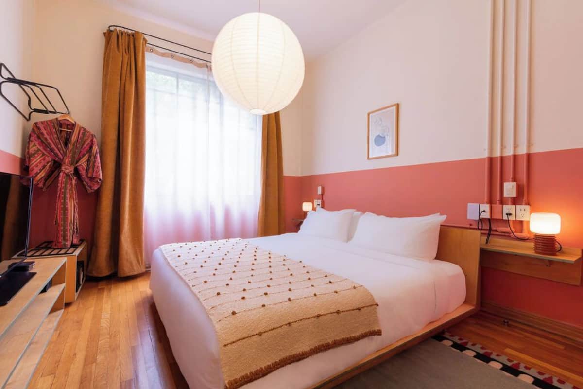 Habitación de hotel con cama, mesita de noche con lámpara, ventana con cortinas y una obra de arte colgada en la pared.