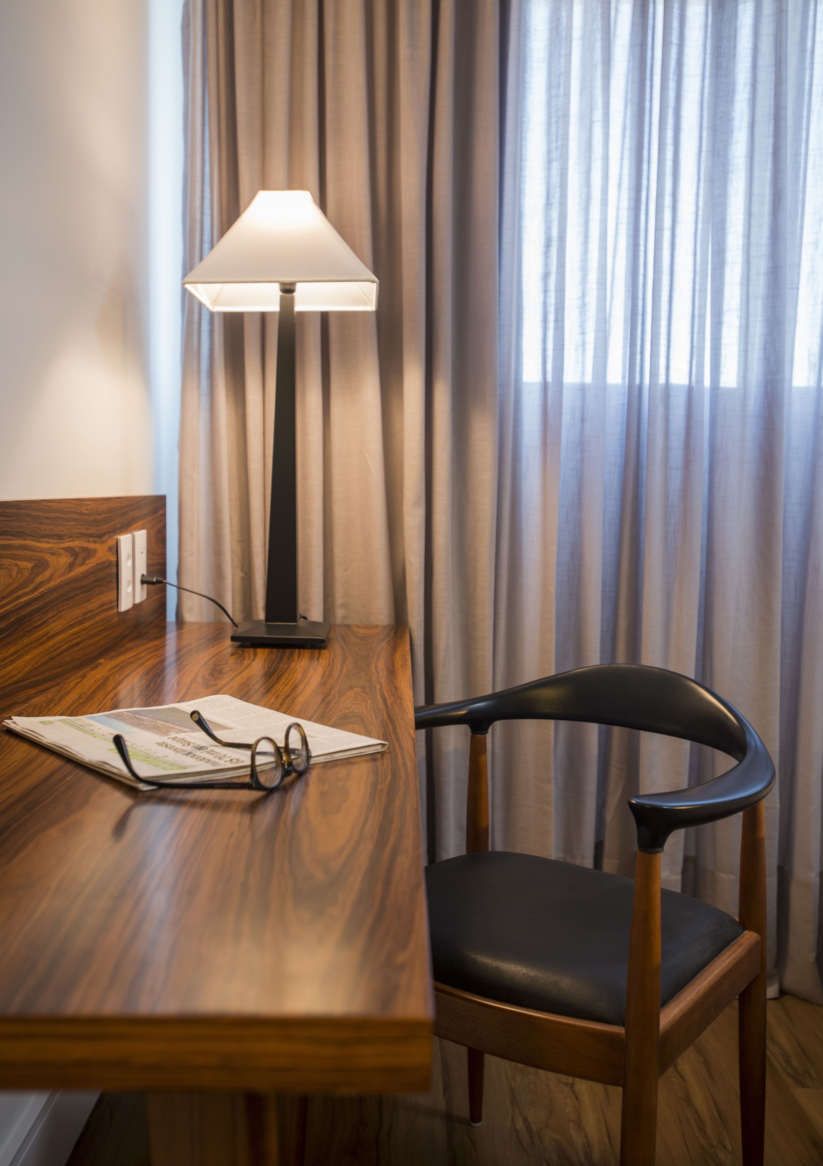 Un escritorio de madera con una lámpara, un par de gafas y una silla en una habitación con cortinas.