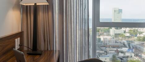 A hotel room with a desk, chair, lamp, and a view of the cityscape through a large window.