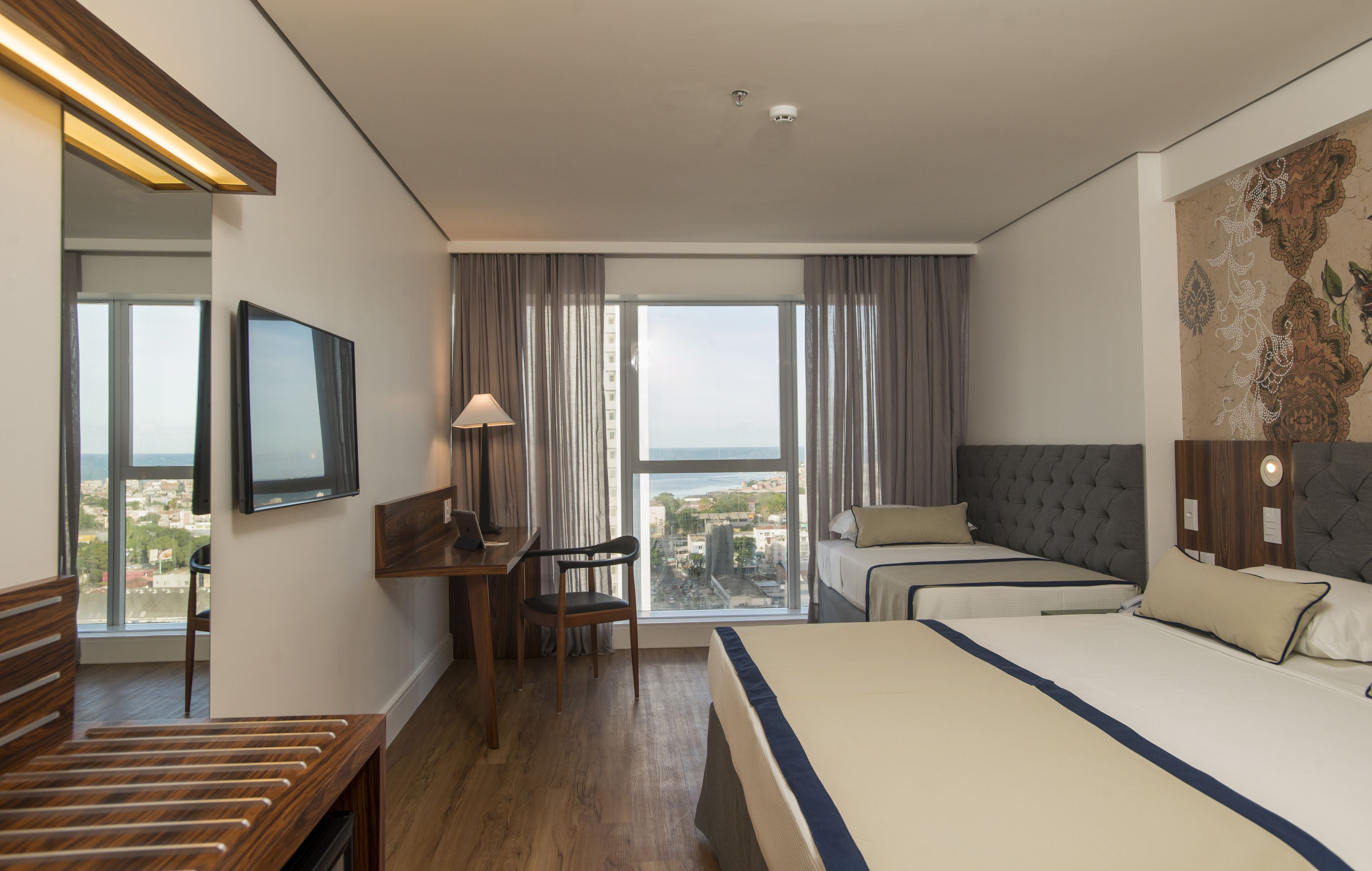 Habitación de hotel con una cama grande, un escritorio con silla, televisor y vistas a la ciudad a través de amplios ventanales.