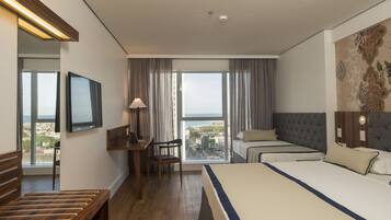 Habitación de hotel con una cama grande, un escritorio con silla, televisor y vistas a la ciudad a través de amplios ventanales.