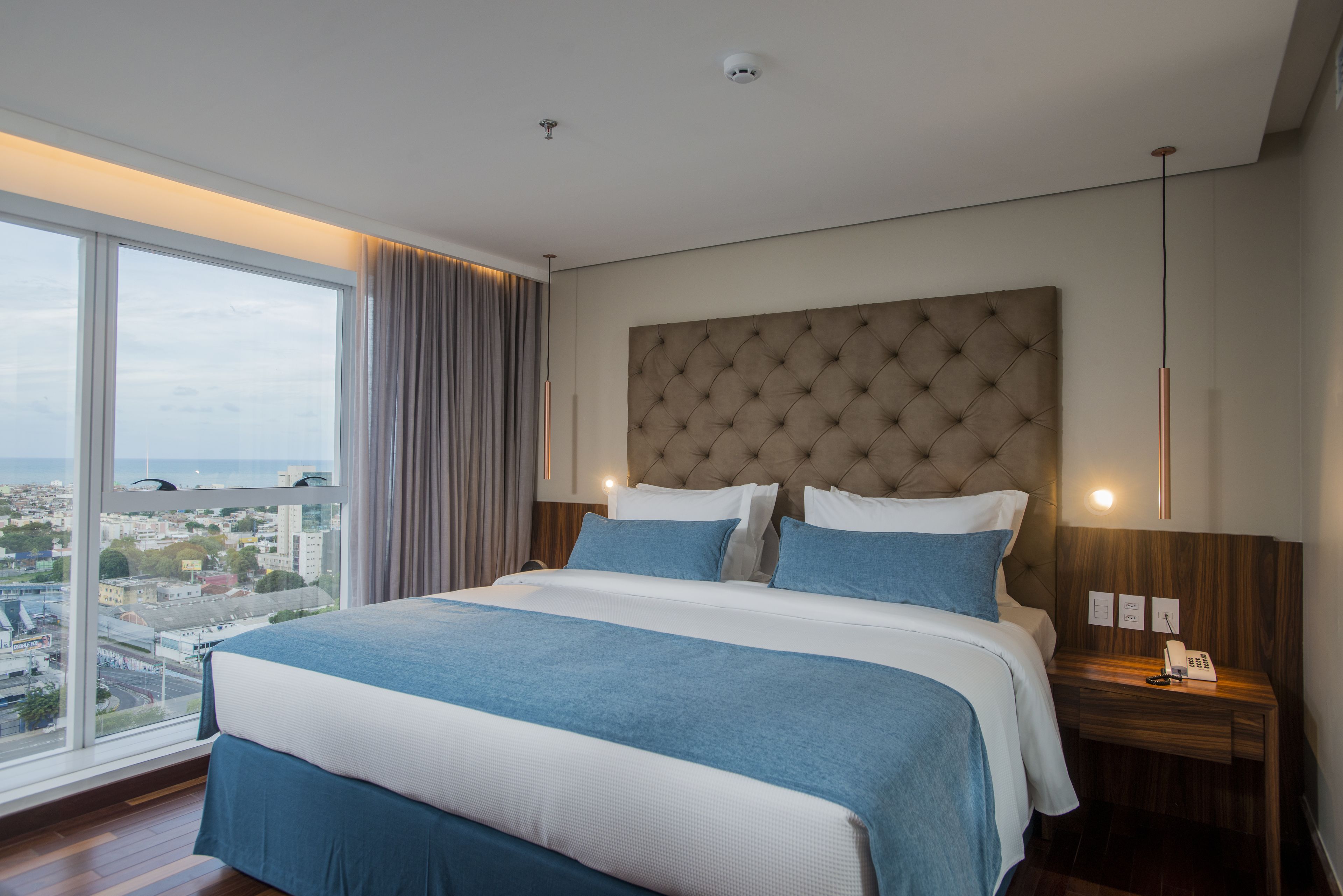 Una habitación de hotel moderna con una cama grande, vistas a la ciudad y una mesita de noche de madera con un teléfono.