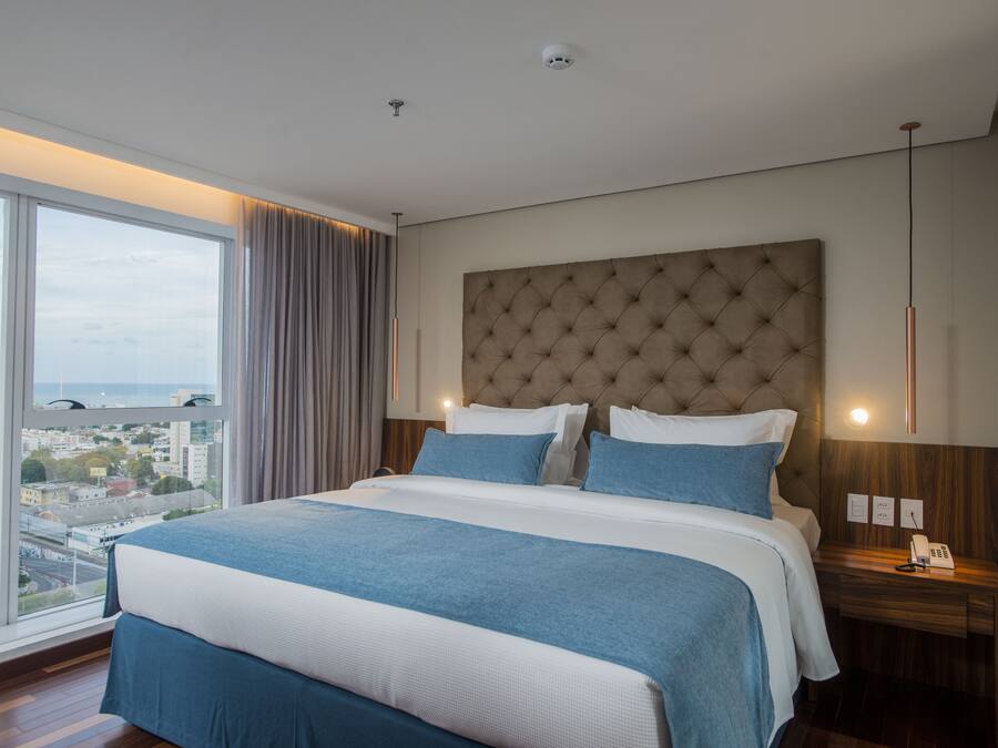 Una habitación de hotel moderna con una cama grande, vistas a la ciudad y una mesita de noche de madera con un teléfono.