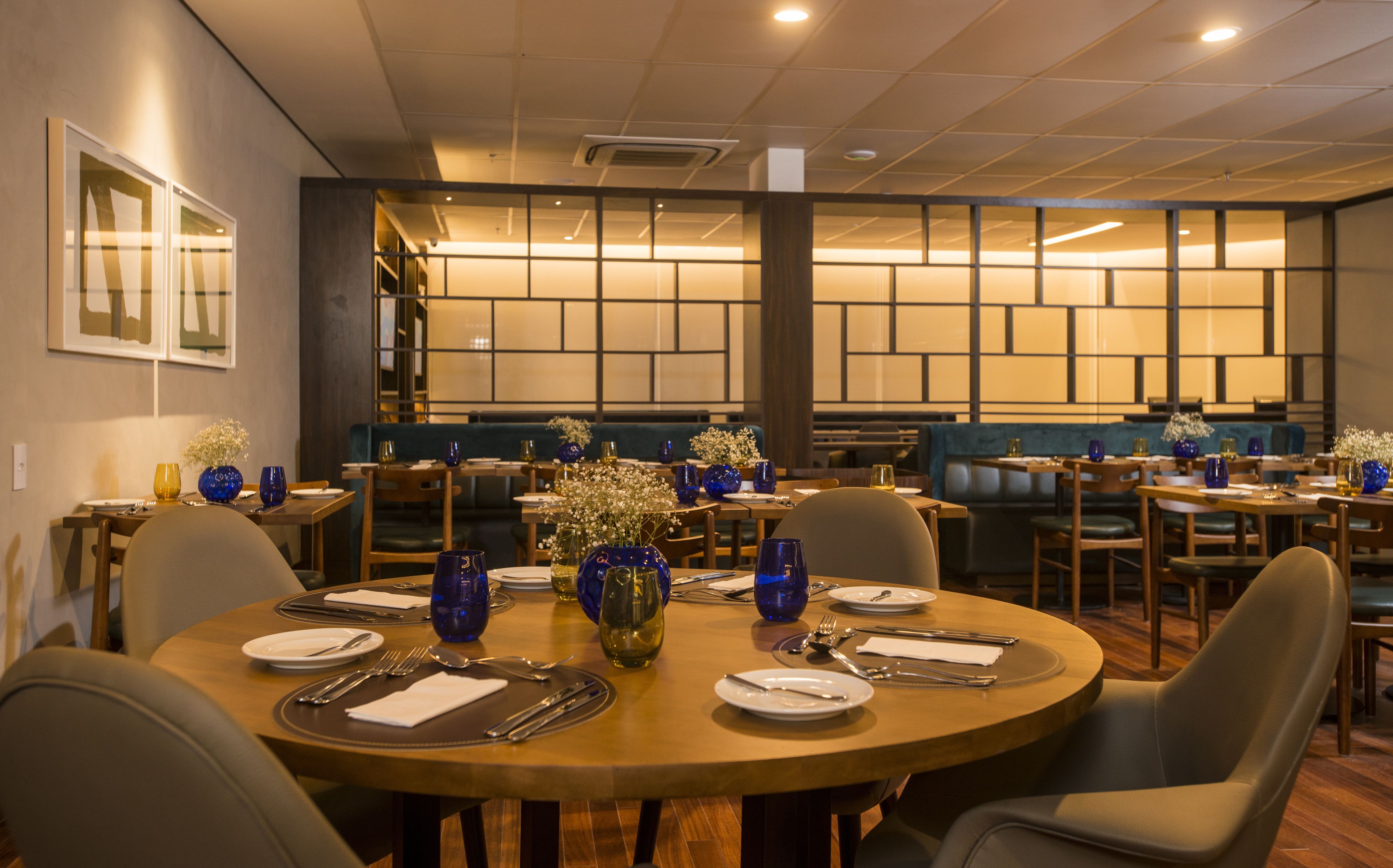 Un restaurante con mesas redondas dispuestas para comer, con vasos de vidrio azul y flores blancas.
