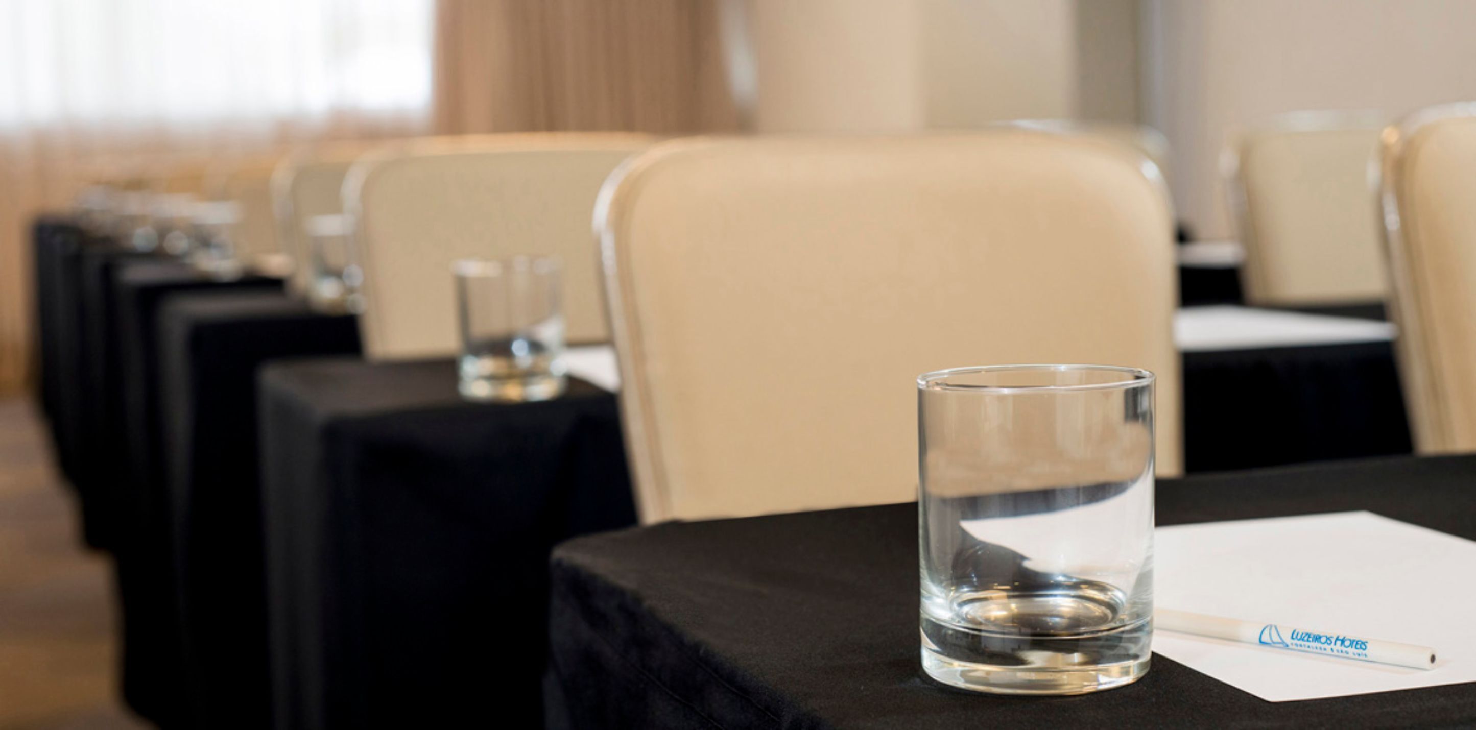 A conference room with rows of beige chairs and black tablecloths, a glass of water, and a pen on a notepad.