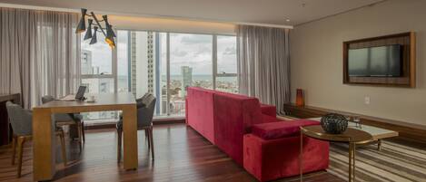 Una sala moderna con un sofá rojo, una mesa de comedor con sillas, un televisor montado en la pared y un ventanal con vistas a la ciudad.