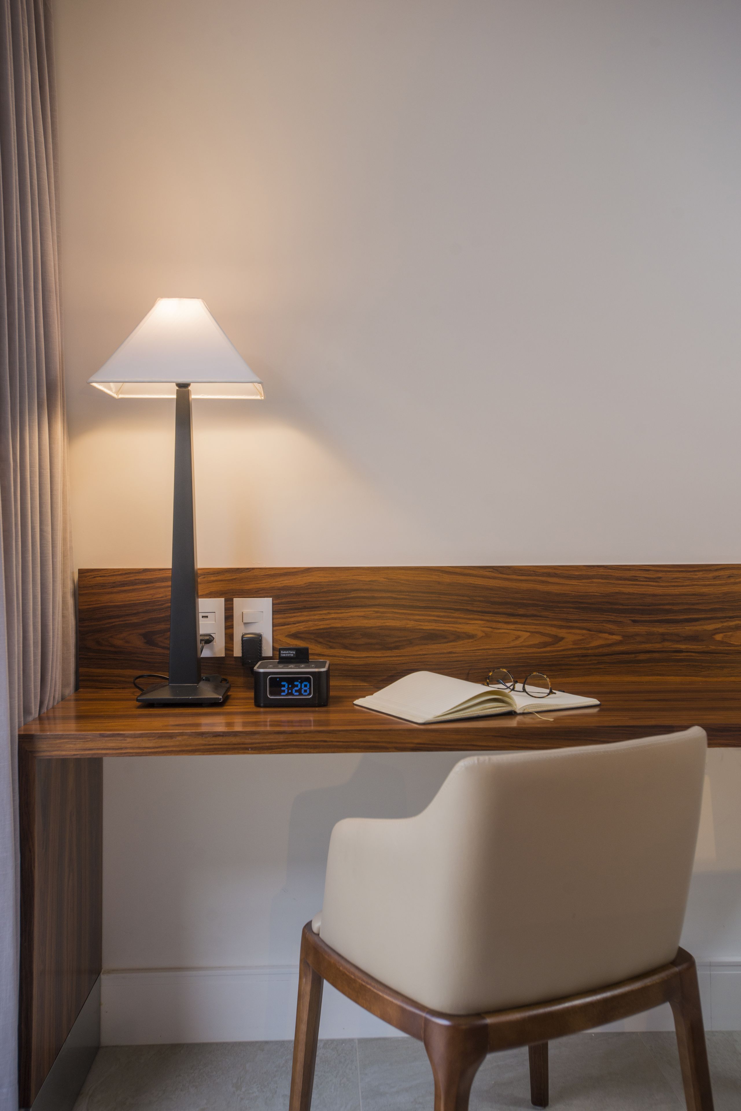 A hotel room with a wooden desk, a lamp, a digital clock, a notebook, and a chair.