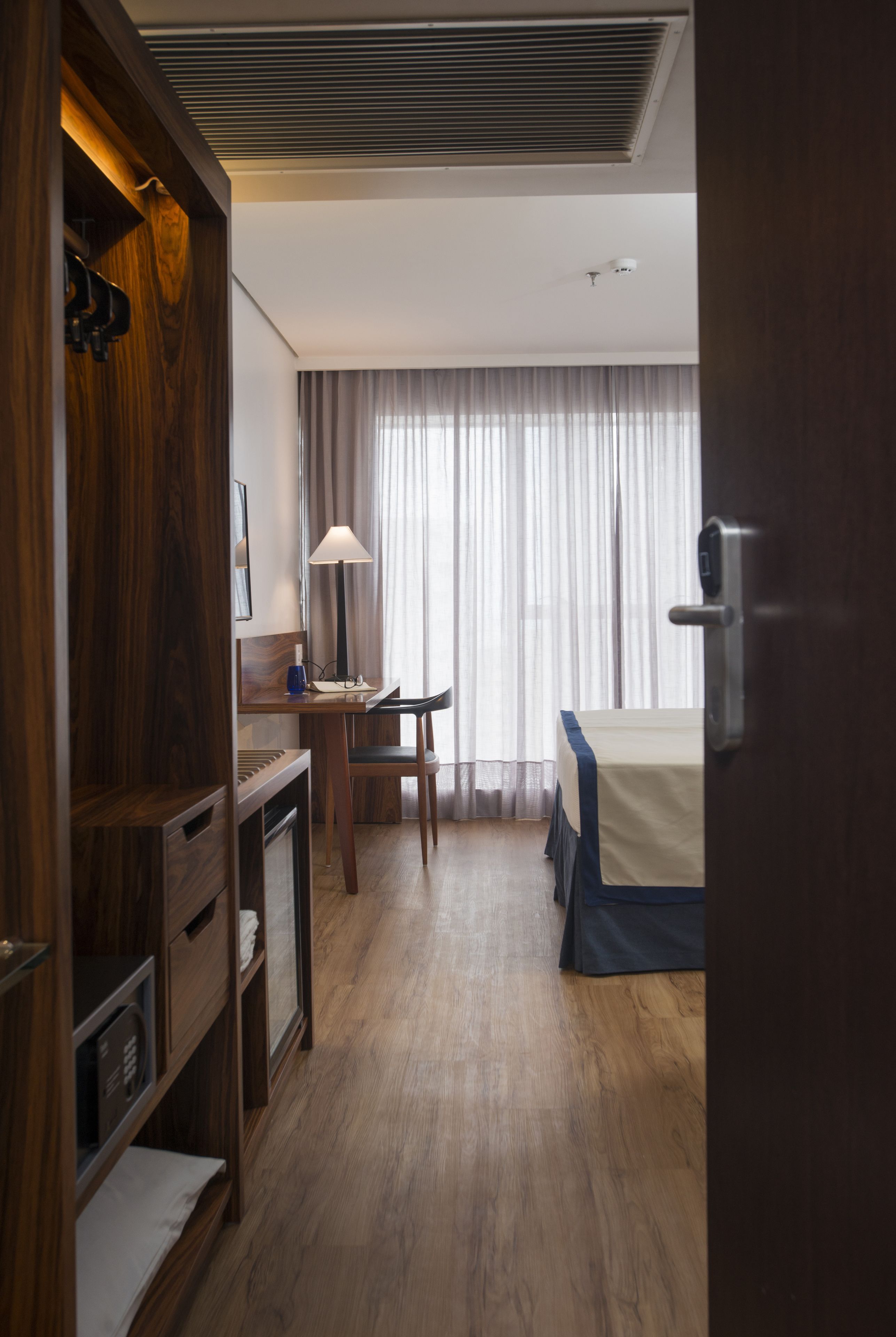 A hotel room with a wooden desk, a chair, a bed with a blue bedspread, a lamp, and a window with curtains.