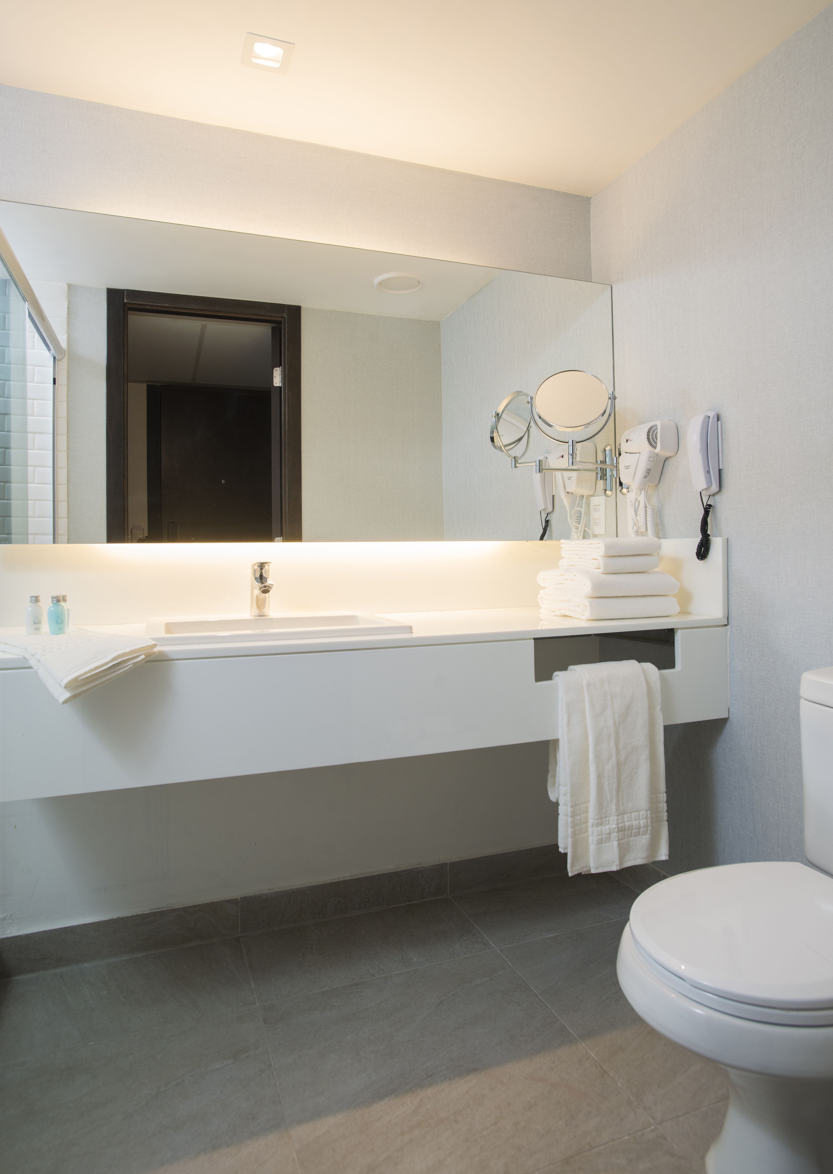 Un baño moderno con lavamanos, espejo y retrete blancos. Hay toallas y artículos de aseo en la encimera.