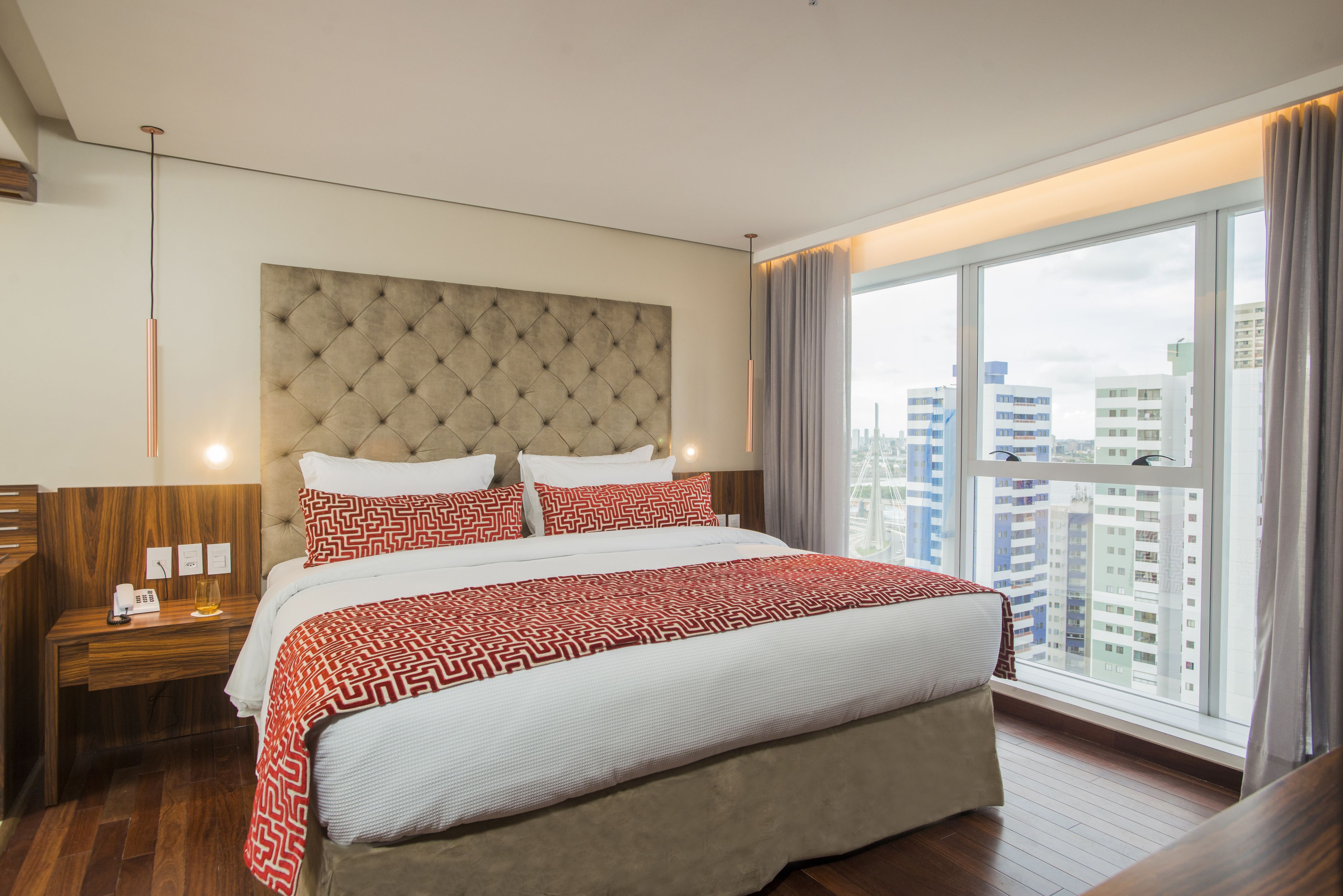 Ein modernes Hotelzimmer mit einem großen Bett, einem Holz-Nachttisch, einem Telefon und Blick auf die Stadt durch große Fenster.
