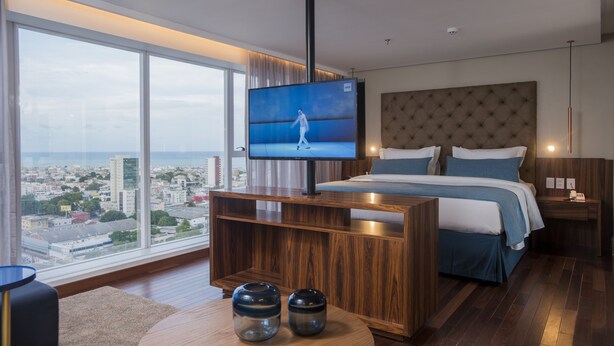 A modern hotel room with a large TV on a wooden console, a bed with blue bedding, a city view, and a round coffee table with two vases. - Hotel Luzeiros Recife (Recife)