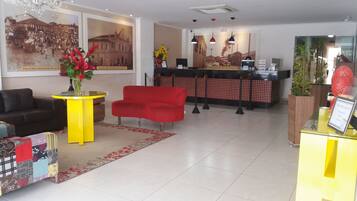 A hotel lobby with a red sofa, a yellow side table with a vase of flowers, a reception desk, and a large framed picture on the wall.