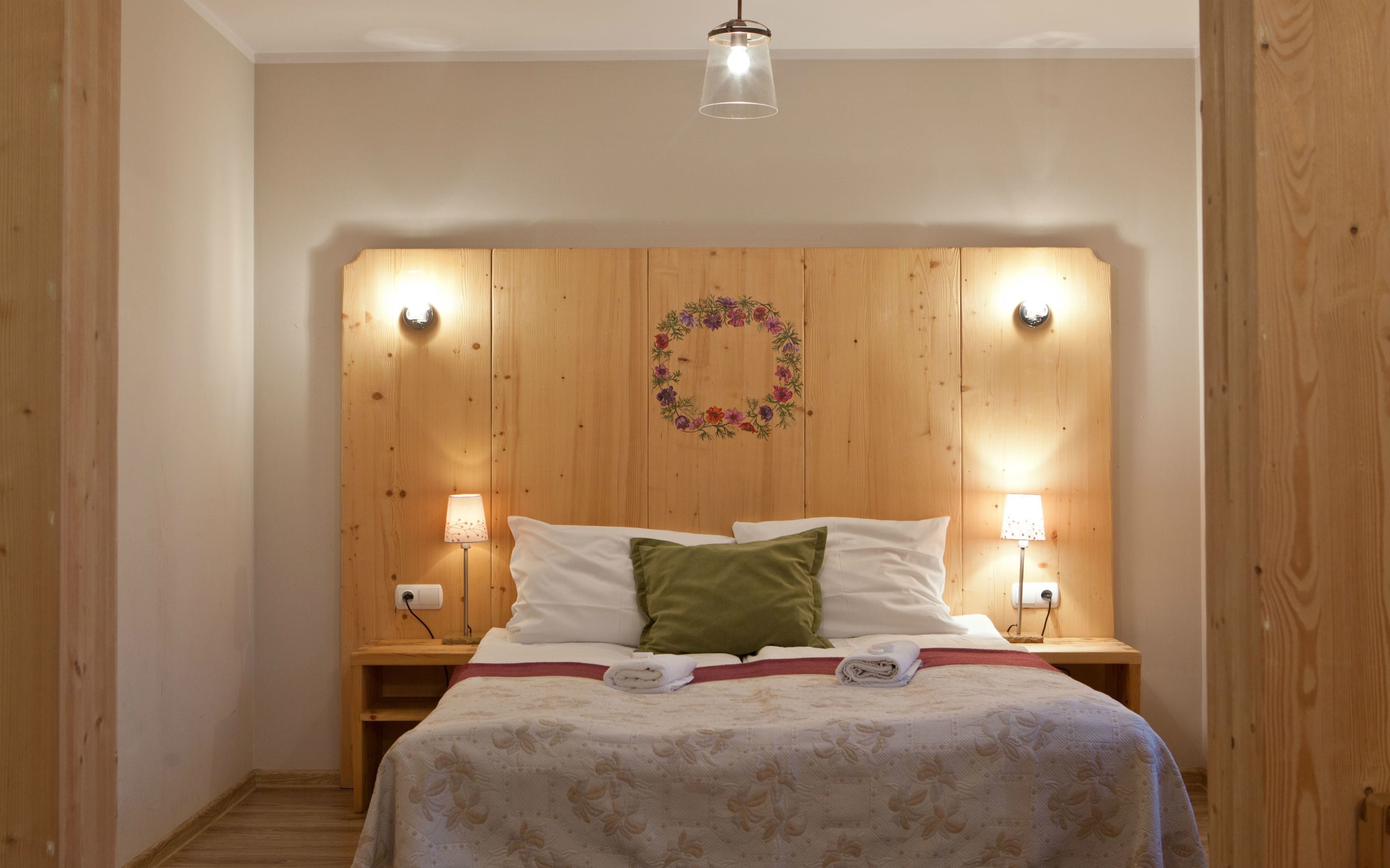 Une chambre à coucher avec une tête de lit en bois, deux lampes de chevet, un lit recouvert d’une couvre-lit à motifs floraux et un luminaire fixé au mur.