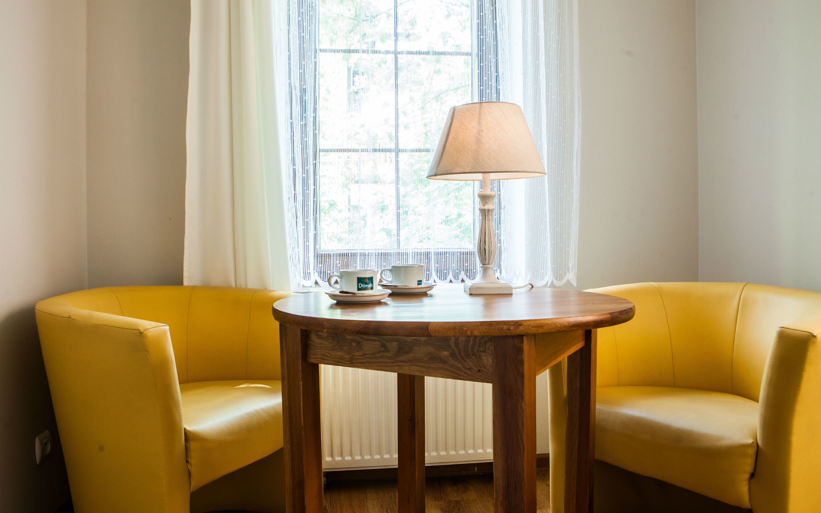 A room with two yellow armchairs, a wooden table with a lamp, and a window with sheer curtains.