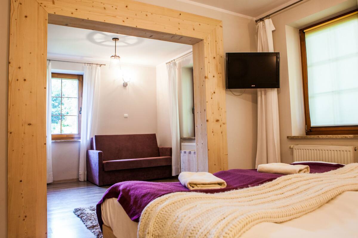 Ein Schlafzimmer mit Bett, Fernseher, Fenster mit Vorhängen und Blick ins Freie.