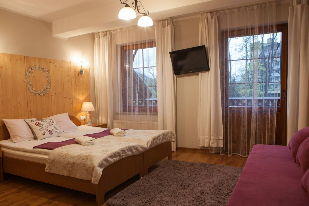 Ein Schlafzimmer mit einem hölzernen Kopfteil, einem Bett mit floralem Bettbezug, einem Nachttisch, einem an der Wand befestigten Fernseher und einem Fenster mit durchsichtigen Vorhängen.