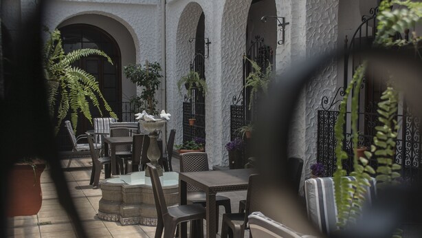 Un patio al aire libre con mesas y sillas, rodeado de plantas en macetas y una pared blanca con arcos.