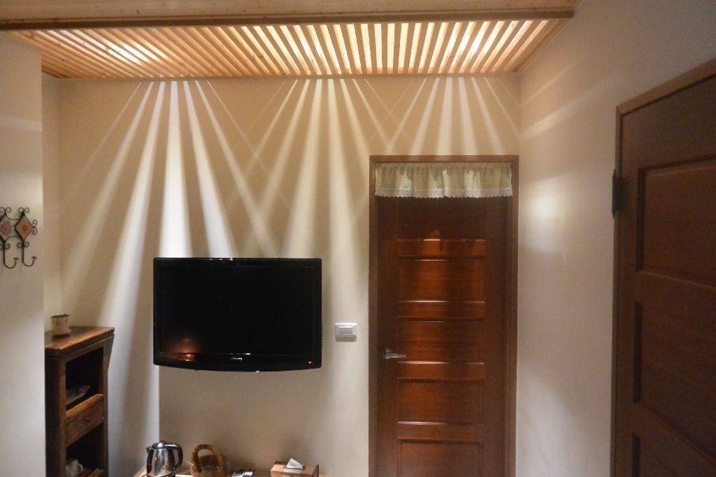 A room with a television mounted on the wall, a wooden door, and a ceiling with slatted beams.