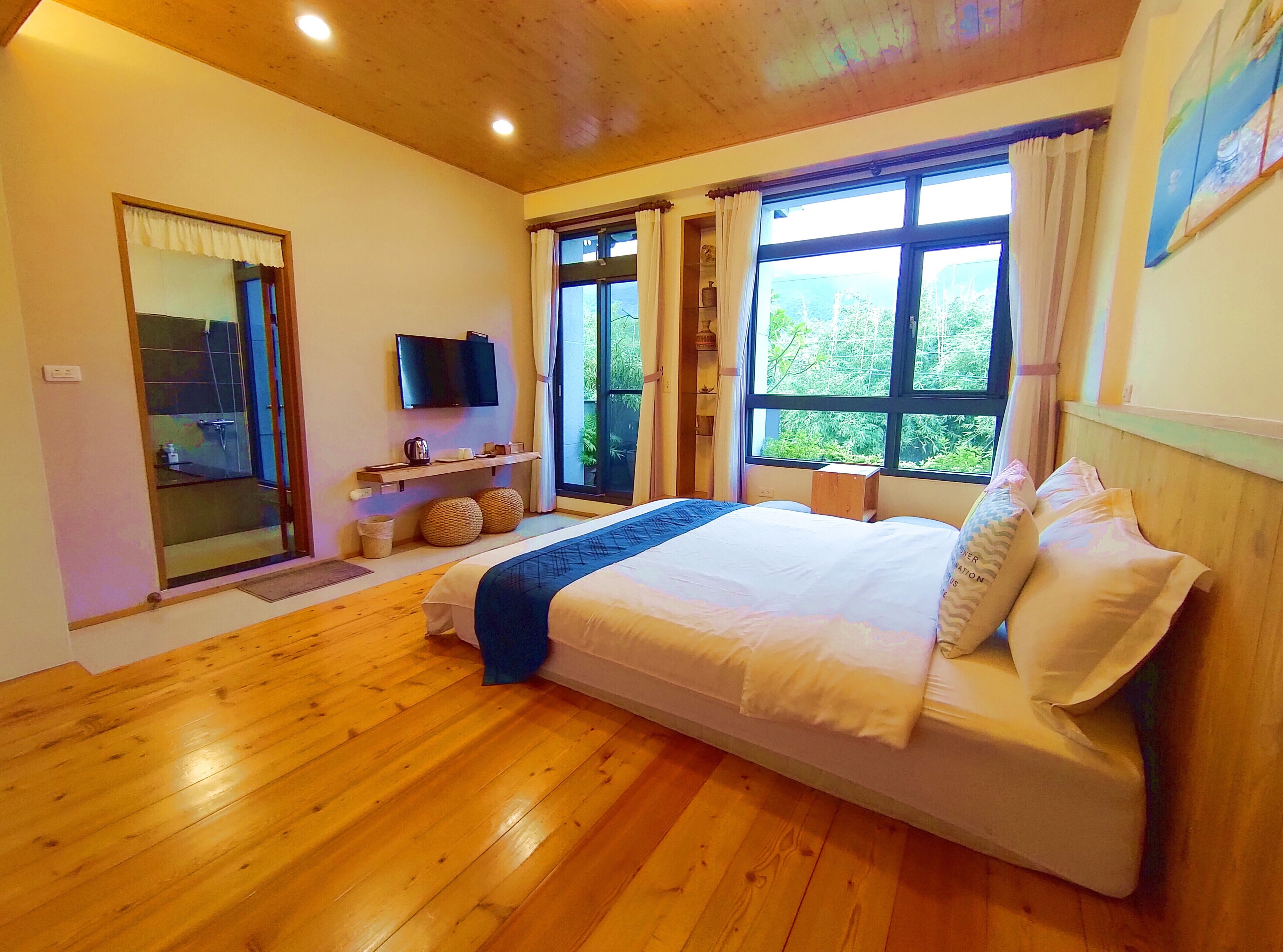 A bedroom with a large bed, wooden flooring, a TV on the wall, and a window with curtains.