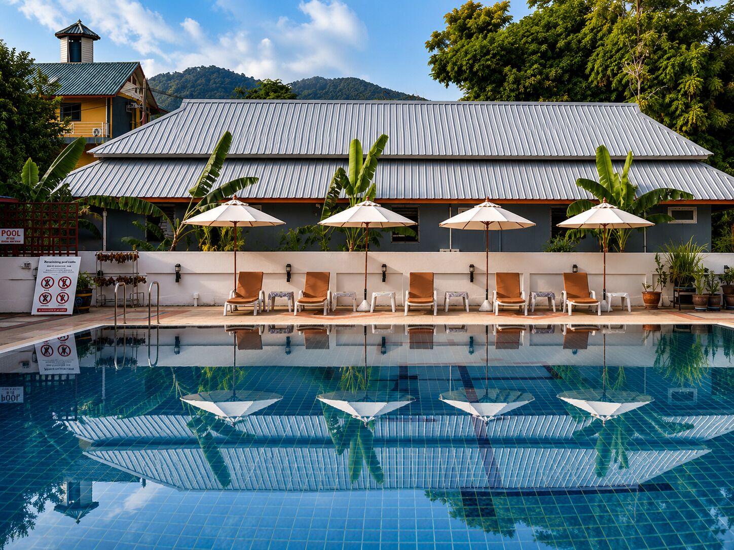 A swimming pool with lounge chairs and umbrellas, surrounded by greenery and a building with a metal roof.
