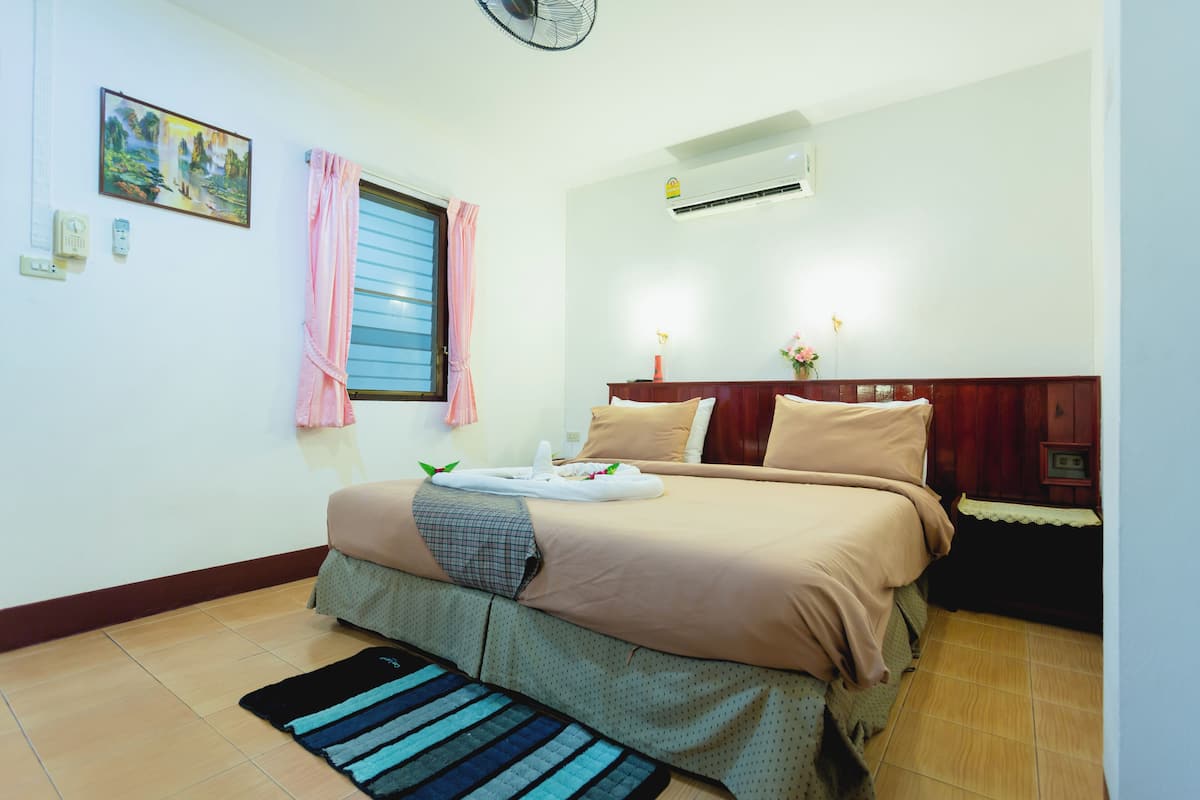 A neatly arranged bedroom with a bed, bedside tables, a window with curtains, an air conditioning unit, and a painting on the wall.