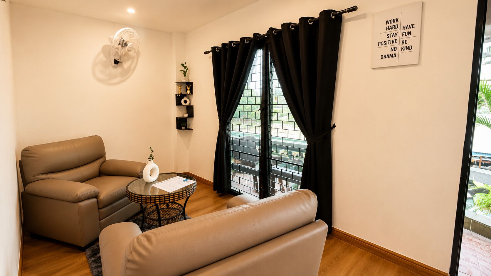 A living room with a sofa, a small round table, a fan, a window with black curtains, and a motivational quote on the wall.