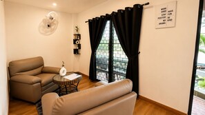 Una sala de estar con un sofá, una mesita redonda pequeña, un ventilador, una ventana con cortinas negras y una frase motivacional en la pared.