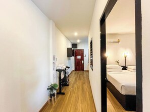 Un pasillo de hotel con piso de madera, una cama con ropa blanca, una planta pequeña y una puerta al final.