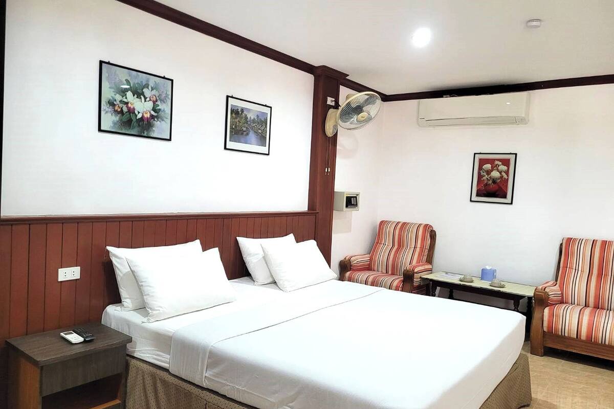 A hotel room with a large bed, two chairs, a small table, and a wall-mounted fan.