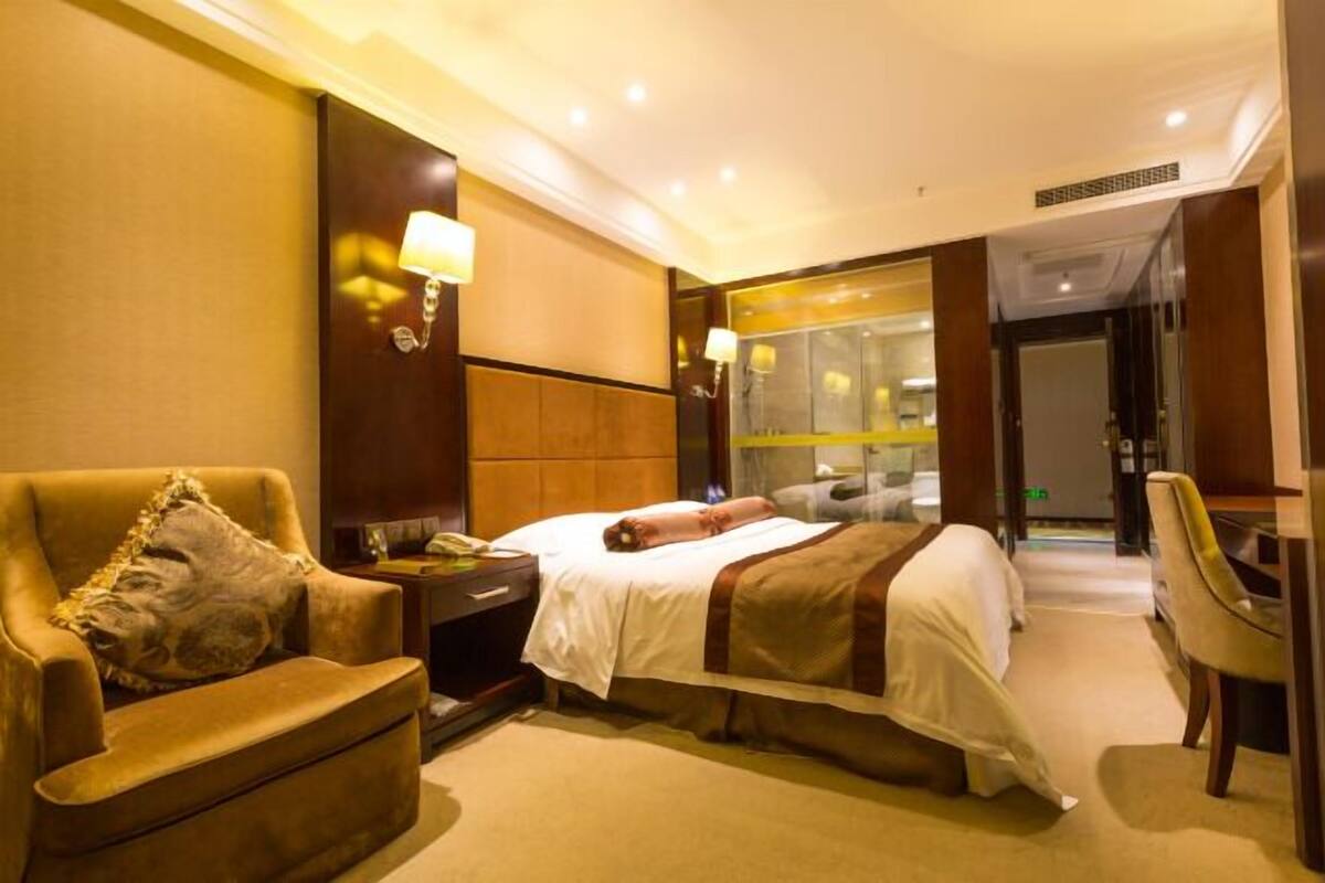 Habitación de hotel con cama, una silla, un escritorio y un baño visible a través de una puerta de vidrio.
